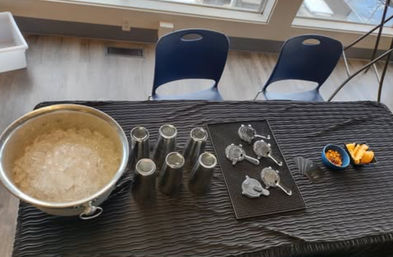Indoor refreshment station by sunlit windows: large ice bucket of crushed ice, six stainless steel cups, cocktail strainers on a black mat, and small bowls of orange slices and snacks on a textured black tablecloth with two blue chairs in the background.