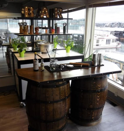 Rustic waterfront indoor bar with a double wooden-barrel counter, cocktail tools and bottles, potted herbs on shelves, and marina view with docked boat