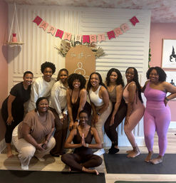Group of women in activewear smiling for a birthday yoga class photo in a pink-themed wellness studio with a 'Happy Birthday' banner, decorative backdrop, and yoga mats — one woman seated in lotus pose with hands in prayer.