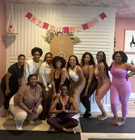 Group of women in activewear smiling for a birthday yoga class photo in a pink-themed wellness studio with a 'Happy Birthday' banner, decorative backdrop, and yoga mats — one woman seated in lotus pose with hands in prayer.