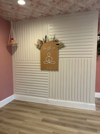 Yoga and wellness studio corner with blush-pink walls, white slatted accent panel, arched tan studio sign with minimalist lotus icon and dried floral arrangement, small hanging shelf with pink pineapple decor, and light wood laminate flooring.