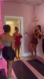 Women in a pink studio standing on yoga mats, one holding a small plate with a lit birthday candle while others cheer, hold drinks and take photos.
