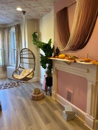 Cozy boho living room with a hanging rattan egg chair, macramé curtains, pink fireplace mantel draped with yarn and small pumpkins, leafy potted plant and wood-look flooring