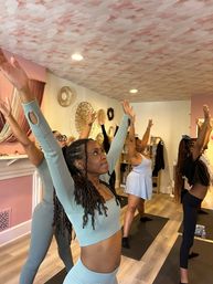 Group yoga class in a bright wellness studio with participants on mats reaching arms overhead in a standing stretch, decorative woven wall art and wood floors.