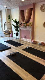 Cozy boho yoga studio interior with black mats and blocks on light wood floors, pink accent fireplace draped with string curtains, hanging rattan swing chair, potted plants and soft cushions.