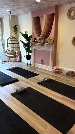 Cozy boho yoga studio interior with black mats and blocks on light wood floors, pink accent fireplace draped with string curtains, hanging rattan swing chair, potted plants and soft cushions.