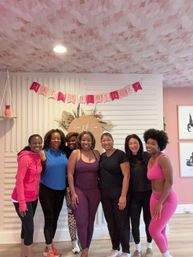 Seven women in colorful activewear posing and smiling in a pink-themed fitness studio for a birthday celebration with a "Happy Birthday" banner and floral backdrop.