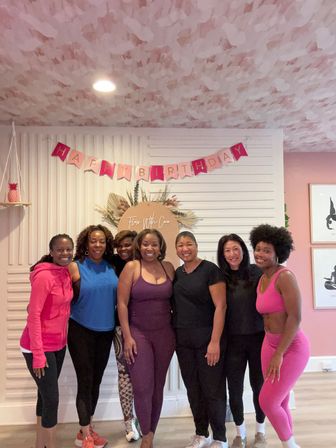 Seven women in colorful activewear posing and smiling in a pink-themed fitness studio for a birthday celebration with a "Happy Birthday" banner and floral backdrop.