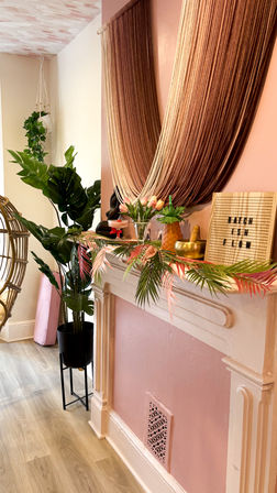 Boho pink interior nook with macramé hanging planter, large tropical potted leaves, draped yarn curtains over a mantel decorated with tulips, brass bowl and a small letter board — cozy feminine decor.