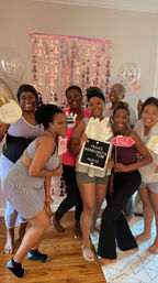 Smiling group of friends at an indoor bachelorette party, holding playful photo props and a letterboard in front of a pink metallic backdrop and balloons in a cozy living room