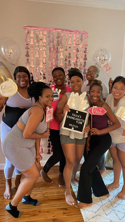 Smiling group of friends at an indoor bachelorette party, holding playful photo props and a letterboard in front of a pink metallic backdrop and balloons in a cozy living room