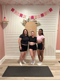 Three smiling women in athletic tops and shorts pose on a yoga mat inside a pink-decorated fitness studio under a "Happy Birthday" banner and dried-floral backdrop.
