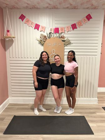 Three smiling women in athletic tops and shorts pose on a yoga mat inside a pink-decorated fitness studio under a "Happy Birthday" banner and dried-floral backdrop.