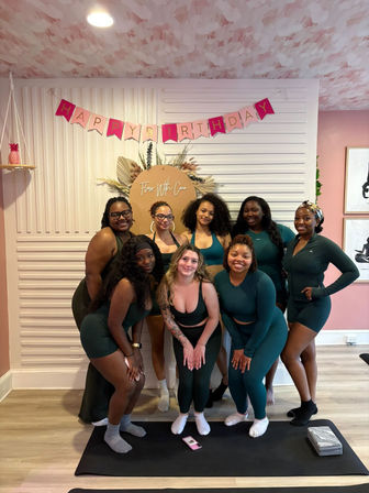 Nine women in matching green activewear smiling together on yoga mats under a pink "Happy Birthday" banner in a bright, pink-accented indoor fitness studio