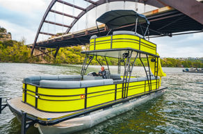 Bright neon-yellow double-decker pontoon boat with canopy on a calm river beneath an arched bridge and treelined shoreline