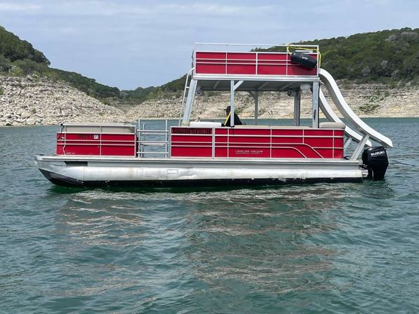 Bright red double-decker pontoon boat with waterslide and outboard motor floating on a clear lake near a rocky shoreline and green hills.