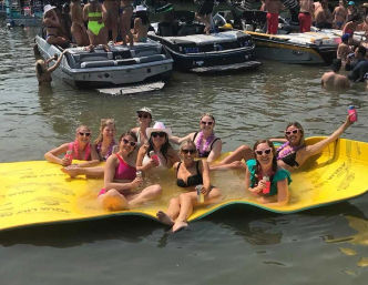 Group of women in swimsuits and playful sunglasses relaxing on a bright yellow floating mat at a crowded lake boat party, holding drinks with powerboats and sunbathers nearby.