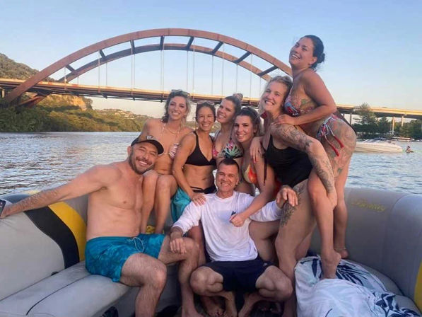 Eight friends laughing and posing on a pontoon boat near a rust-colored arch bridge over a calm river at sunset, summer vibe