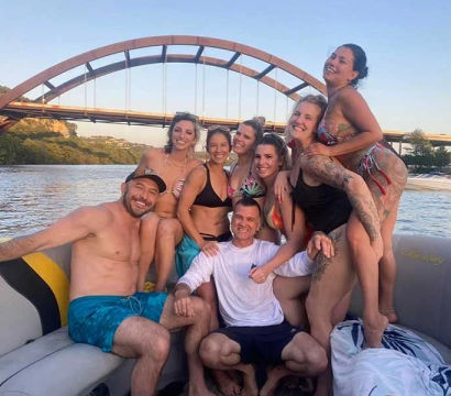 Eight friends laughing and posing on a pontoon boat near a rust-colored arch bridge over a calm river at sunset, summer vibe
