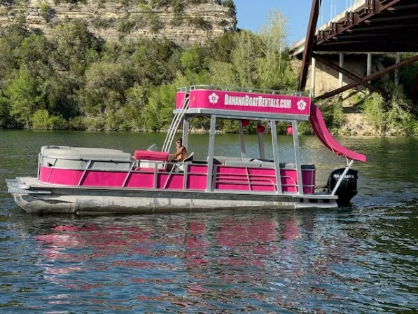 Bright pink pontoon party boat with a waterslide floating on a calm river beneath a steel bridge and rocky cliff, framed by green trees.