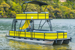Bright yellow two-level pontoon boat with upper deck, canopy and waterslide floating on a calm lake by a green, tree-lined shoreline.