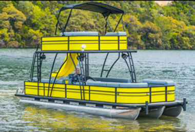 Bright yellow two-level pontoon boat with upper deck, canopy and waterslide floating on a calm lake by a green, tree-lined shoreline.