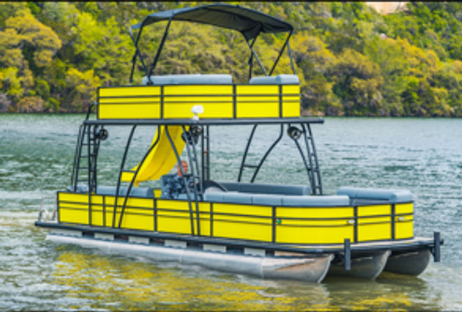 Bright yellow two-level pontoon boat with upper deck, canopy and waterslide floating on a calm lake by a green, tree-lined shoreline.