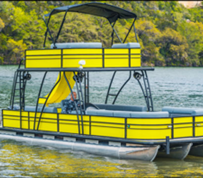 Bright yellow two-level pontoon boat with upper deck, canopy and waterslide floating on a calm lake by a green, tree-lined shoreline.