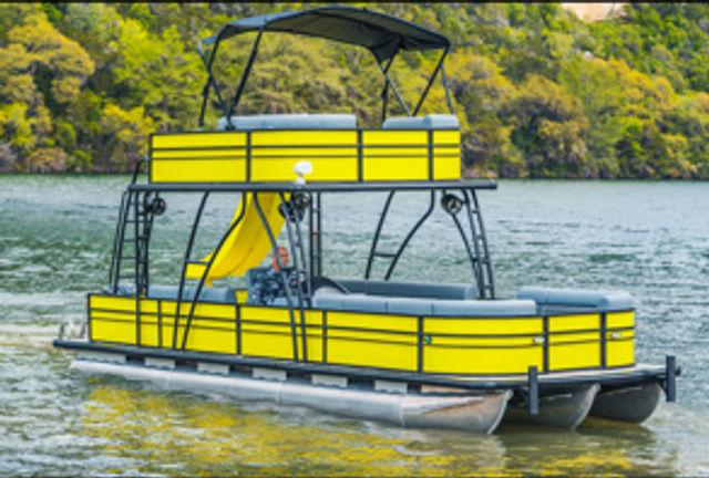 Bright yellow two-level pontoon boat with upper deck, canopy and waterslide floating on a calm lake by a green, tree-lined shoreline.