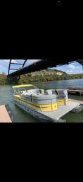 Cheerful yellow-and-gray pontoon boat docked at a wooden pier beneath an arched steel bridge on a calm river with tree-covered bluffs and blue sky.