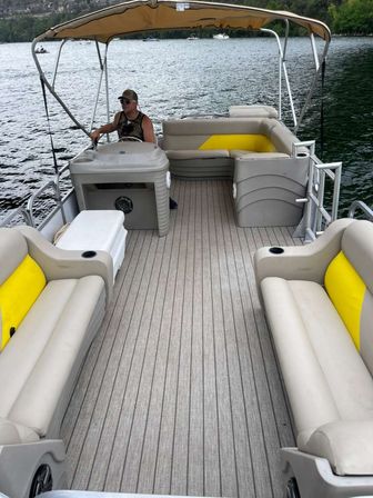 Pontoon boat interior with beige and yellow vinyl benches, teak-look flooring and a tan canopy, a person at the helm steering on a calm, tree-lined lake.