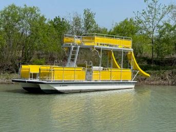 Bright yellow double-decker pontoon boat with waterslide and upper deck floating on a calm river beside a leafy green riverbank