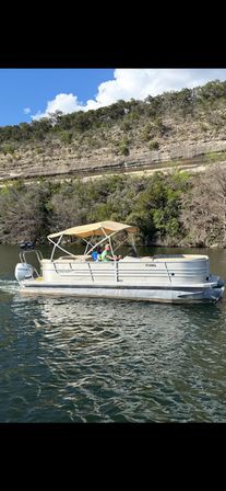 Pontoon boat with tan canopy carrying two people cruising on a calm lake beneath layered limestone cliffs and oak-covered hills under a bright blue sky.