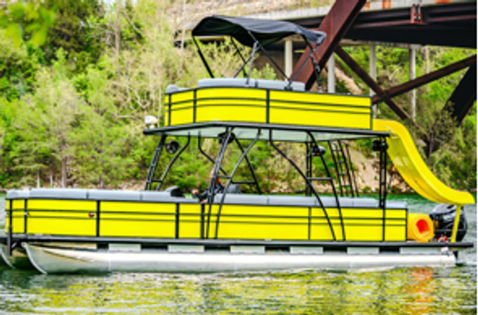 Bright yellow pontoon boat with a curved waterslide moored under a bridge on a calm, tree-lined lake