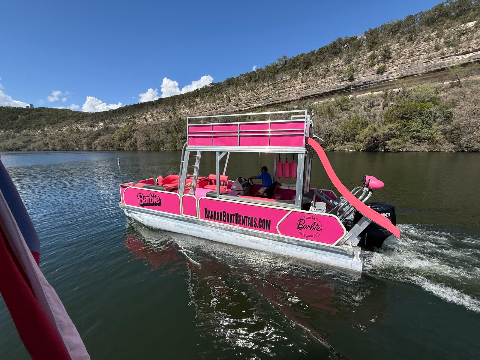Bright pink party pontoon boat with upper deck and curved slide cruising on a cliff-lined lake beneath a clear blue sky