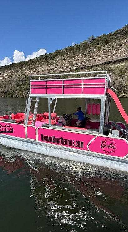Bright pink party pontoon boat with upper deck and curved slide cruising on a cliff-lined lake beneath a clear blue sky