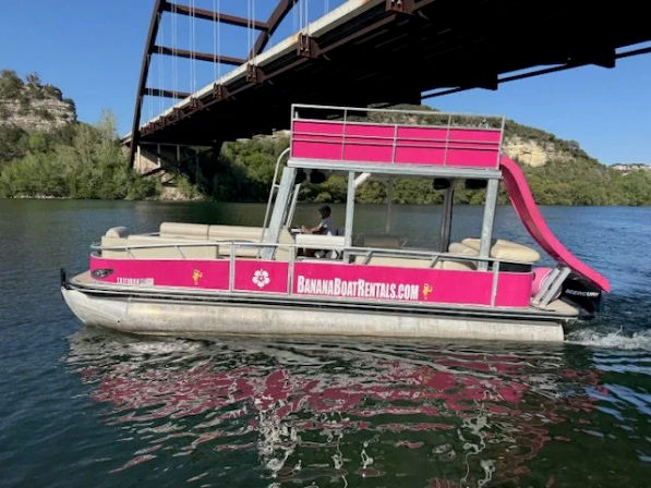 Bright pink pontoon rental boat with rooftop waterslide and driver cruising a calm river under a steel arch bridge, sunlight reflecting on the water.