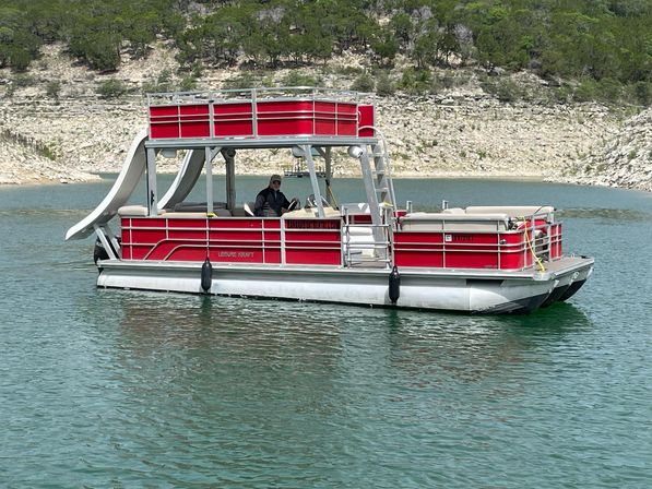 Red double-decker pontoon boat with waterslide and upper deck, person at the helm, cruising on a turquoise lake near a rocky, tree-lined shoreline