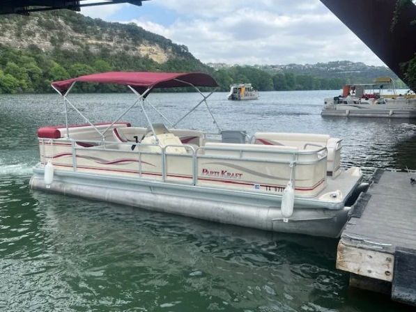 Pontoon boat with maroon canopy docked at a lakeside pier, calm green water with tree-lined shore and limestone bluffs under a cloudy sky.