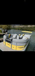 Yellow-and-gray pontoon boat with curved vinyl seating and matching canopy docked at a wooden pier on a calm lake, tree-lined shoreline and rocky bluff in the background — ready to cruise.