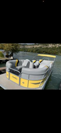 Yellow-and-gray pontoon boat with curved vinyl seating and matching canopy docked at a wooden pier on a calm lake, tree-lined shoreline and rocky bluff in the background — ready to cruise.