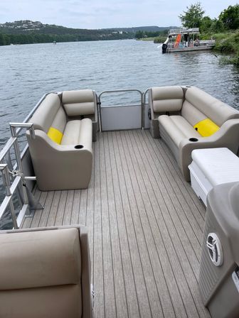View from a pontoon boat's rear deck with beige bench seating and yellow cushions, looking across a calm lake toward a tree-lined shoreline, distant hills and other boats docked nearby.