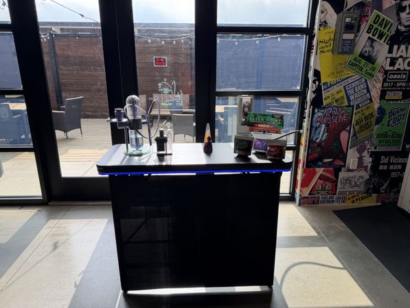 Sunlit indoor hookah station on a counter with a glass water pipe and product packs, set before large glass doors leading to a wooden outdoor patio with wicker chairs and a colorful music-poster wall.