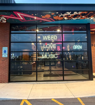 Evening brick storefront with glass garage-style windows and neon signs reading "WEED LOVE MUSIC" and "OPEN", colorful mural overhang and reserved accessible parking in front.