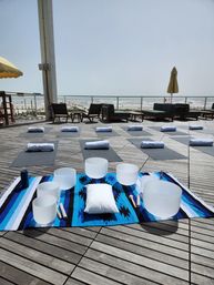 Oceanview beachfront deck set for a coastal sound‑bath yoga session — rows of yoga mats with rolled towels and crystal singing bowls on a blue blanket.