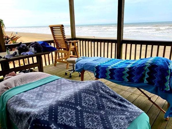 Two massage tables draped with colorful patterned blankets on a wooden beachfront deck overlooking a sandy shore and ocean waves, with a wooden chair and small table of supplies nearby.