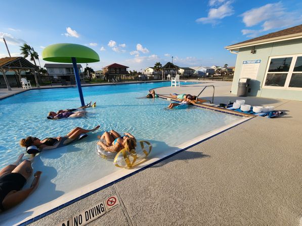 People lounging on floaties in a shallow outdoor pool with a green mushroom water feature, palm trees and coastal neighborhood homes under a sunny blue sky