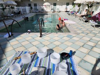Sunny hotel pool deck with pale blue tiled floor, guests lounging and swimming, and crystal singing bowls on a striped blanket for a poolside sound bath