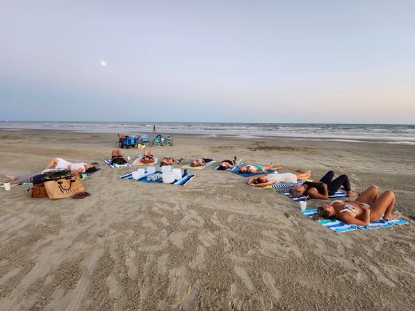 Group of people relaxing on striped beach towels in a loose semicircle on a wide sandy beach at dusk, facing the sky near the ocean with a visible moon and white bowls and chairs nearby — relaxed coastal sunset gathering.