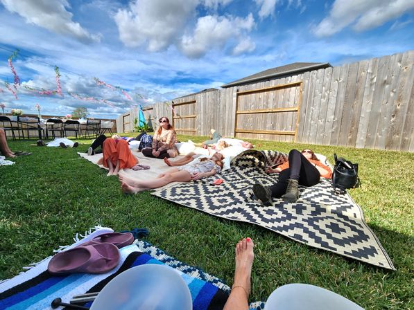 Group of people lounging on patterned rugs and blankets in a sunny backyard picnic setting with green grass, a wooden fence, festive hanging garlands, flip‑flops and a bright blue sky with clouds.